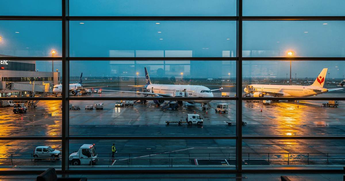 Вид из окна терминала на мокрый перрон в сумерках: три самолёта у телетрапов, багажные тележки и оранжевые огни фонарей