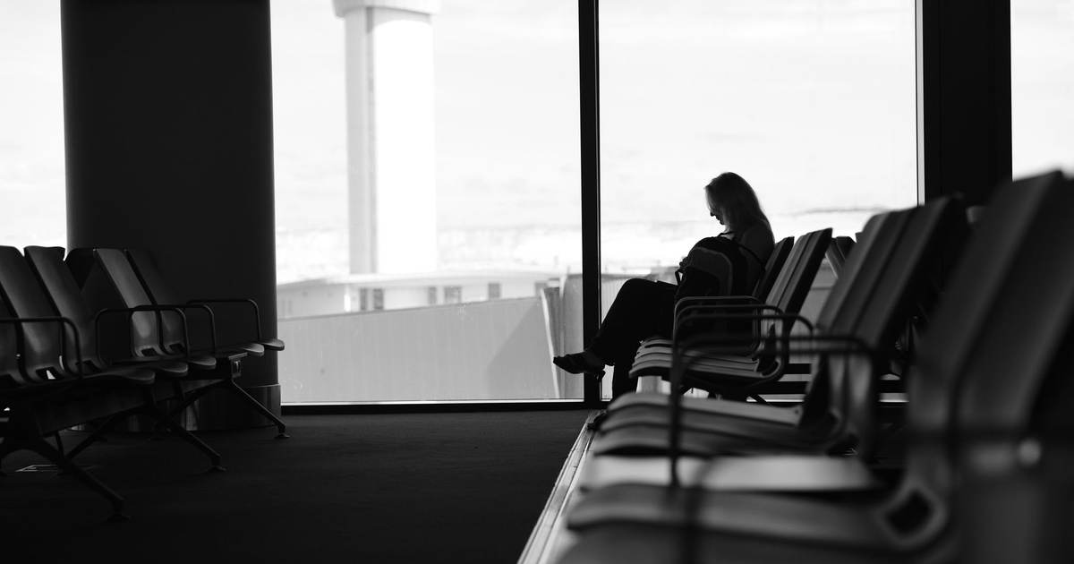 Силуэт человека в кресле у окна зала ожидания, за стеклом пасмурное небо и диспетчерская вышка, чёрно-белый кадр